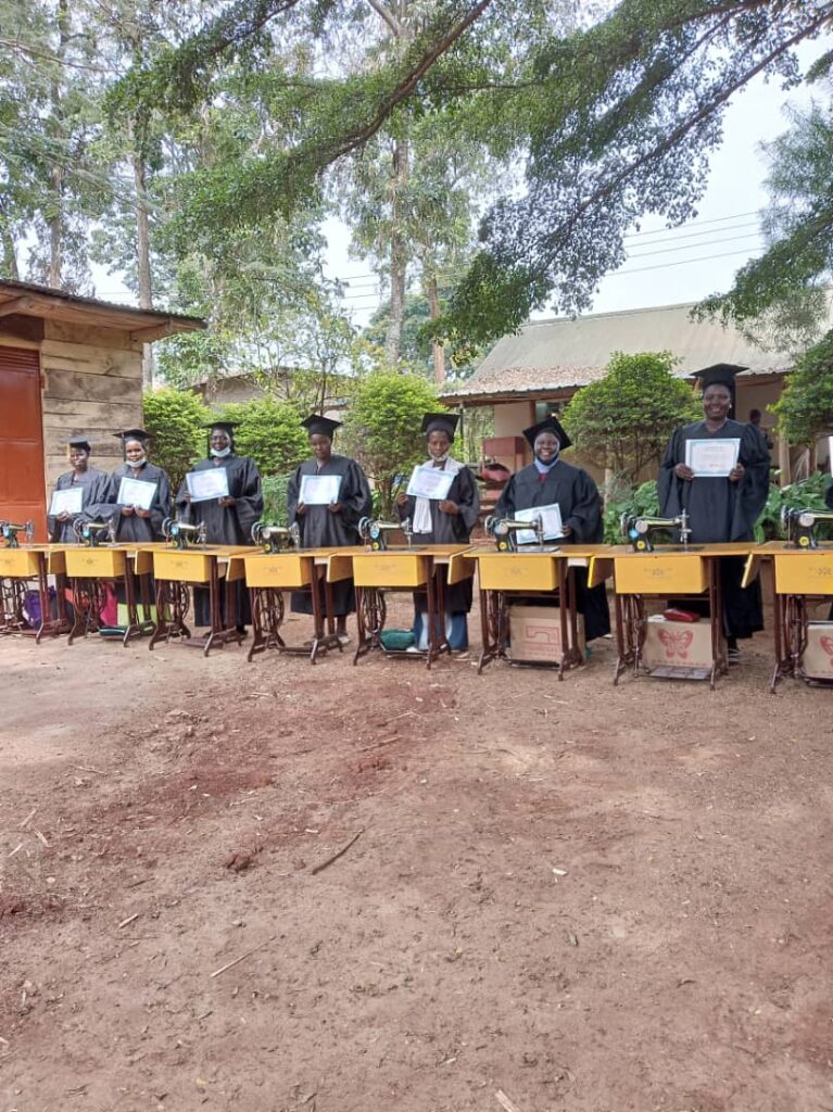 Lynette and friends wearing their graduation gowns and holding their certificates. On the desks in front of each person is a sewing machine