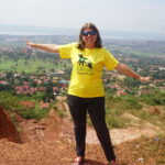 Nicola on a hill top in Uganda, with her arms out smiling. It is a sunny day and Nicola is wearing a SALVE t-shirt, looking happy.