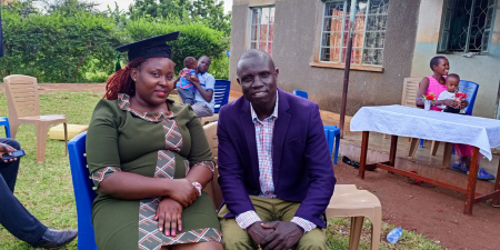 A young woman is wearing a graduation cap and gown, posing for the camera sat next to a man.