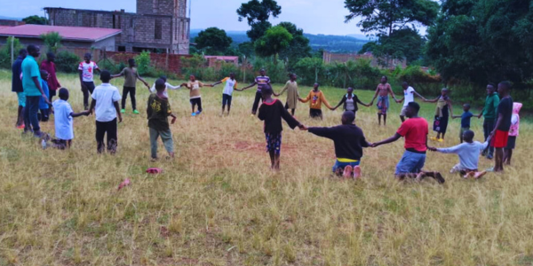 A group of children are standing in a large circle, holding hands in a field.