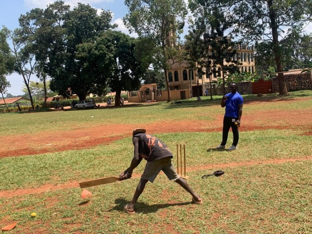 A boy is playing cricket while a coach looks on. The boy is standing in front of the wickets holding a bat.